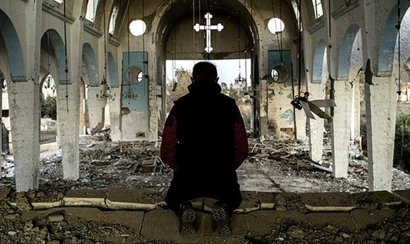 Líder cristão se ajoelha sob destroços de igreja destruída no Oriente Médio. (Foto: Reuters)