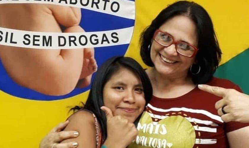 Damares Alves (direita) e Lulu (esquerda) abraçadas em frente à bandeira de uma campanha contra o aborto. (Foto: Instagram)