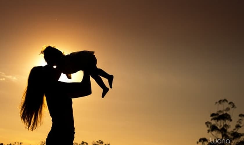 Mãe levanta a filha nos braços durante um por do sol. (Foto: Luana Okaeda)