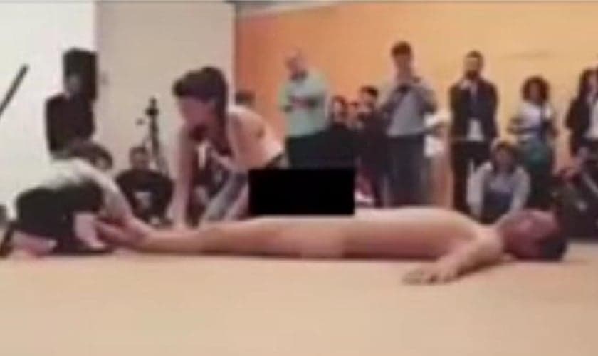 Criança de cerca de quatro anos (esquerda) é estimulada por mulher adulta (centro) a tocar corpo nu do homem que está deitado no tablado durante performance do '35º Panorama da Arte Brasileira – 2017'. (Imagem: Reprodução - Internet)